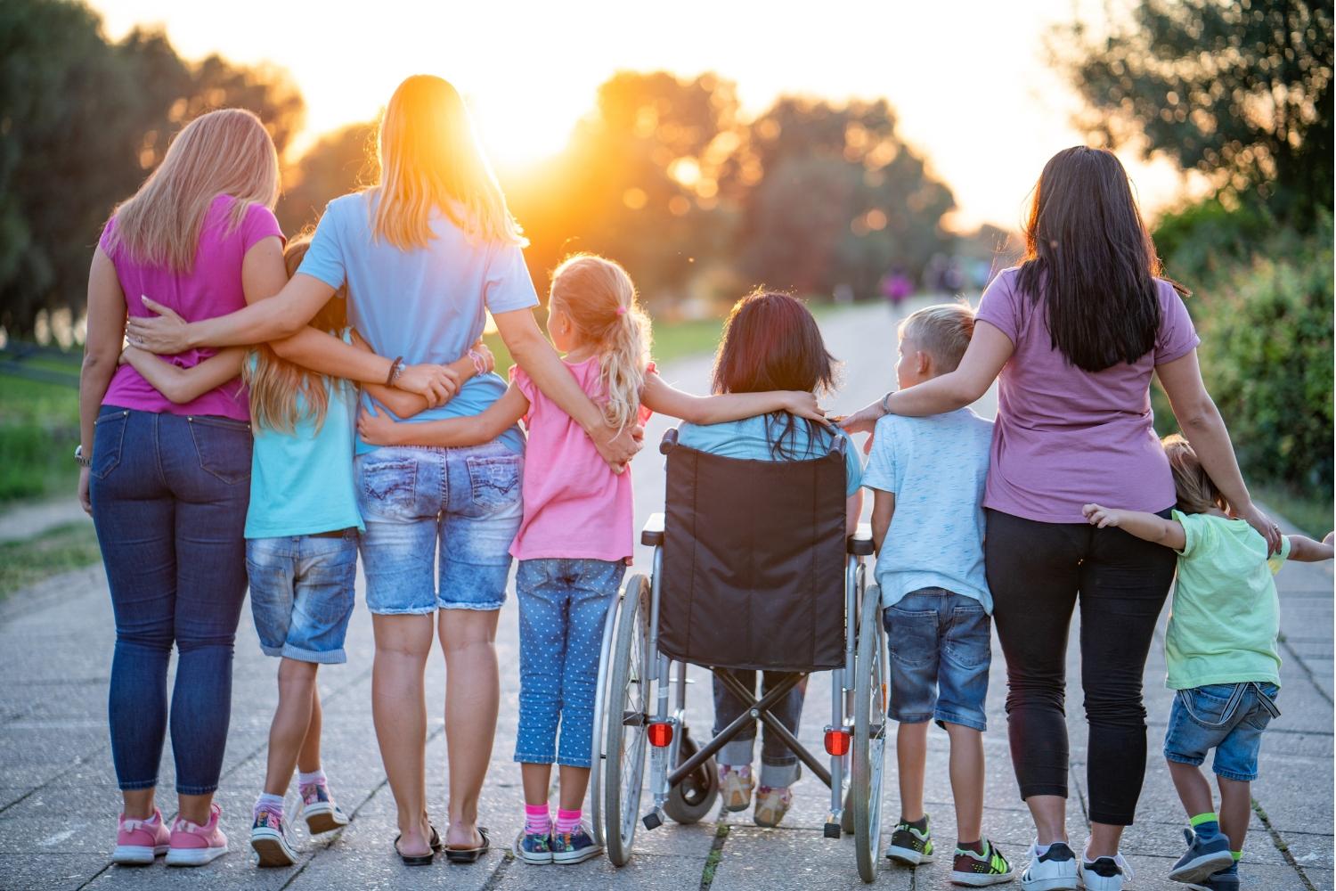 Diverse group of adults and children in a line are standing and sitting in wheelchair with their arms around each other, with their backs towards the camera facing the sunset.