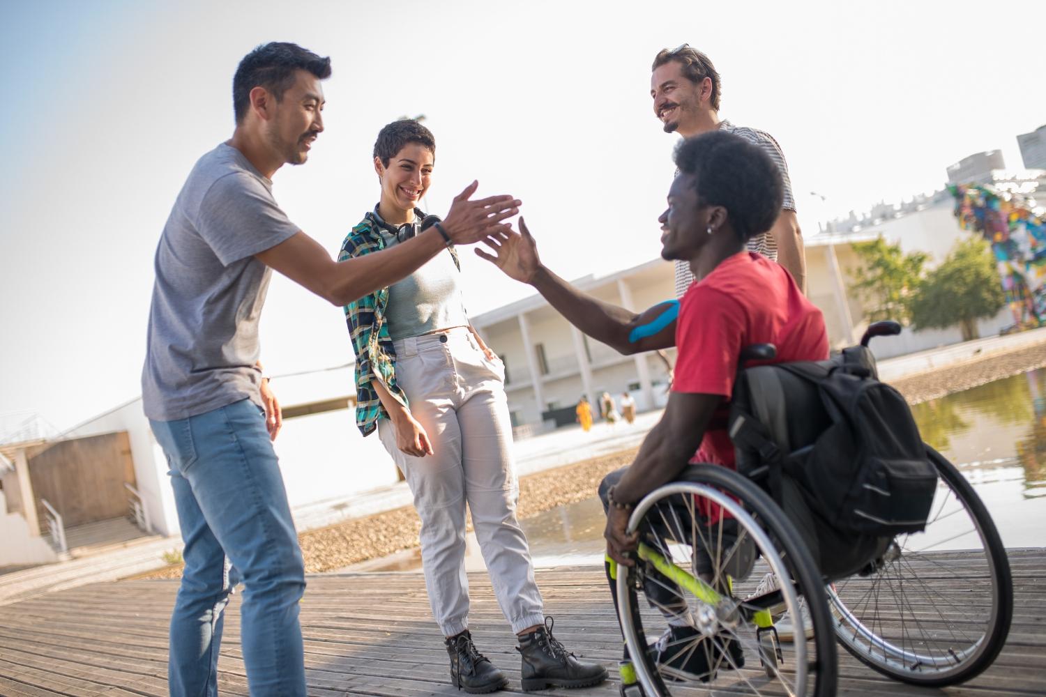 A group of four friends, including a person in a wheelchair, smile and greet each other with high-fives while standing and talking outdoors near a waterfront on a sunny day.