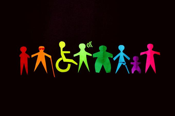 Colorful paper cutouts of diverse human figures, including representations of people with disabilities, a child, and various body types, stand side by side against a black background, symbolizing inclusion and diversity